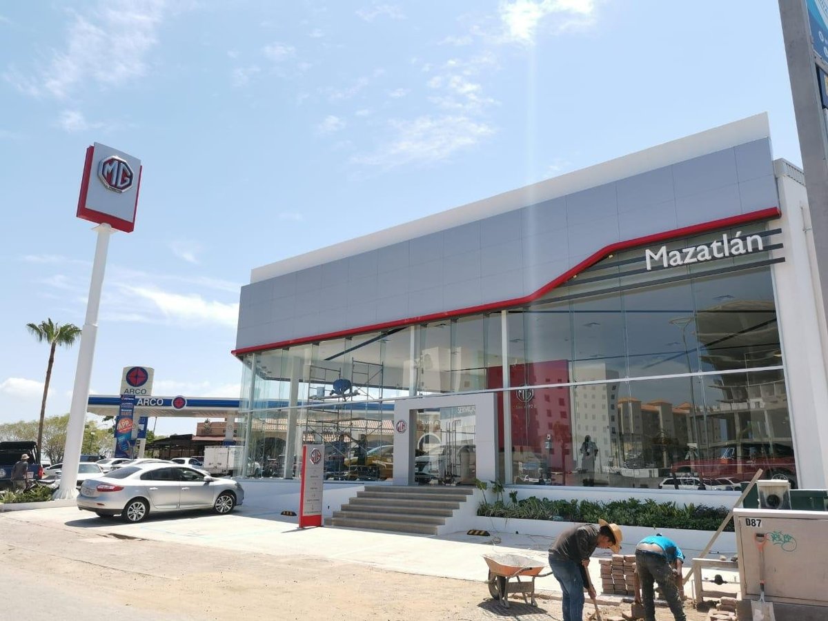 MG motor dealership storefront with red signage, modern architecture, and parking lot with vehicles on a sunny day